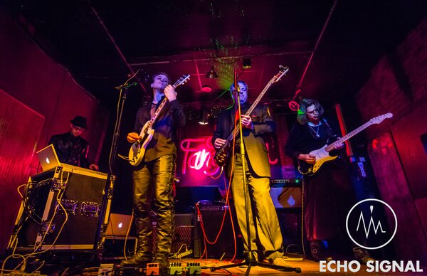 Charles, Adam, John, and Phaedrus (Echo Signal in 2016) playing live at the Red Sea, the stage washed in red with a partially obscured Fangtasia neon sign behind the band.